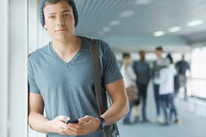 Junger Mann mit Mütze und T-Shirt in gleicher Farbe steht an eine Wand gelehnt und guckt kurz von seinem Smartphone hoch