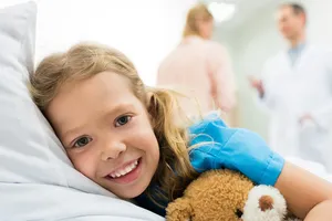 Kleines Mädchen mit Teddy im Arm dreht sich im Bett um und von den beiden Pflegekräften im Raum weg