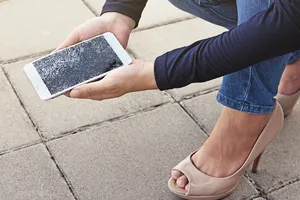 Junge Frau kniet auf der Straße und hält fassungslos ihr Smartphone mit dem gesprungenen Display in der Hand