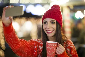 Junge Frau schießt mit rotem Glühweinbecher in der Hand ein Selfie vor der heimeligen Kulisse des örtlichen Weihnachtsmarktes