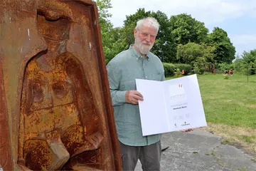Der Preisträger des Kunstpreises 2021 Reinhard Buch posiert mit der Urkunde neben einem seiner Werke