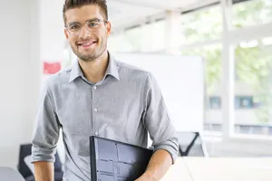 Junger Mann in hellgrauem Hemd mit hochgekrempelten Ärmeln sitzt mit seiner Präsentationsmappe halb auf dem Tisch und lächelt vorsichtig