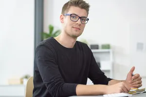 Junger Mann in dunklem Pulli guckt von seinem vor ihm liegenden Laptop hoch und lächelt leicht