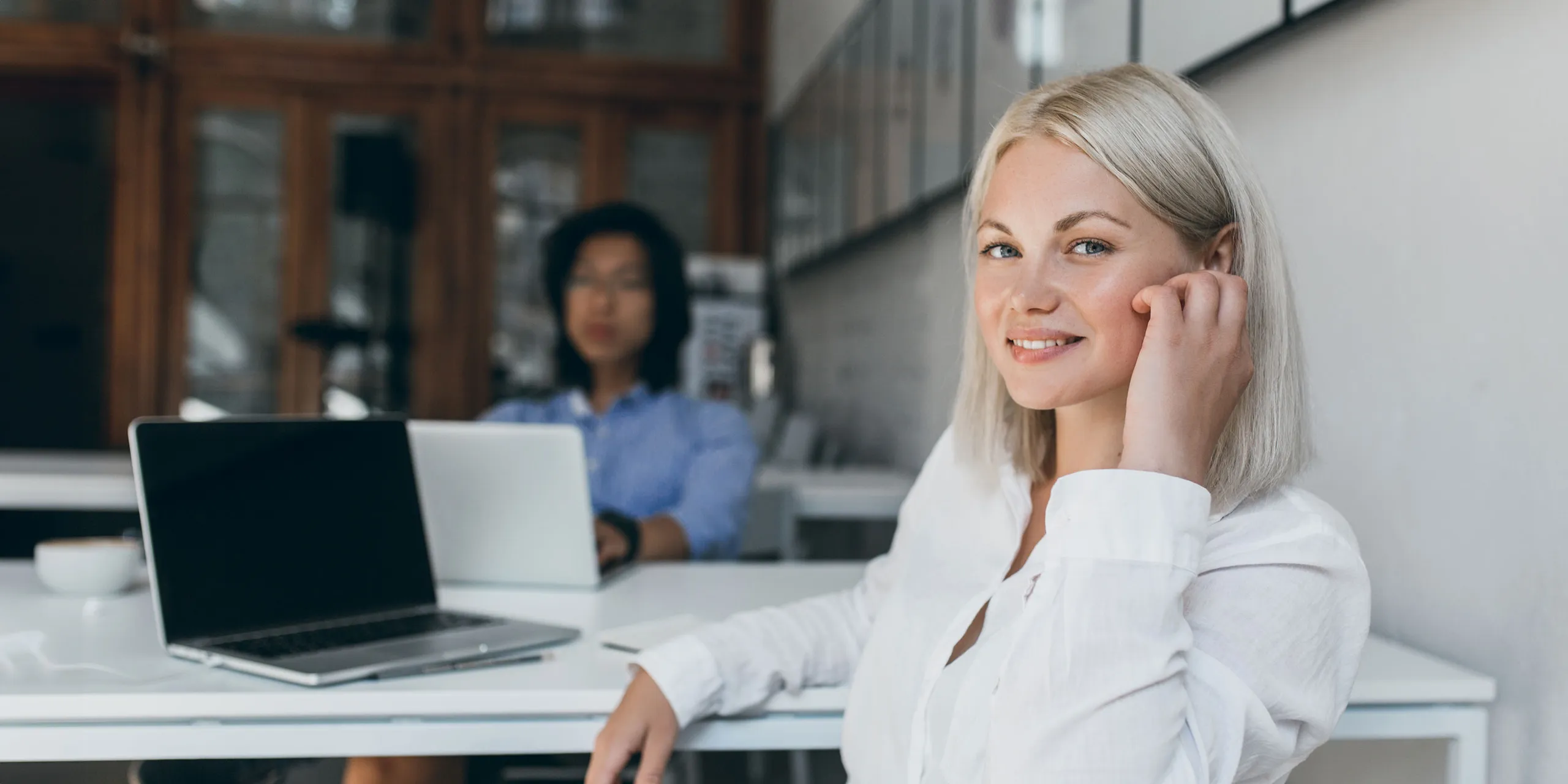 Junge Frau mit blonden Haaren und weißer Bluse sitzt lächelnd an einem Schreibtisch mit Laptop