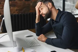 Mann im dunklen Hemd sitzt gestresst am Schreibtisch vor einem Computer und hält sich den Kopf