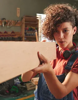 Junge Frau mit brauen Locken und Latzhose vermisst ein Holzbrett in einer Werkstatt