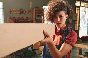 Junge Frau mit brauen Locken und Latzhose vermisst ein Holzbrett in einer Werkstatt