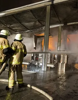 Zwei Feuerwehrleute löschen noch die letzten Glutnester nach einem Großbrand in einer Lagerhalle